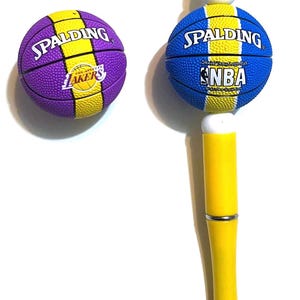 May include: Two basketball-themed pens. One pen has a purple and yellow basketball with the Los Angeles Lakers logo. The other pen has a blue and yellow basketball with the NBA logo. Both pens have yellow barrels.