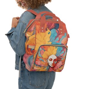 May include: A red and orange backpack with a colorful abstract design featuring a woman's face. The backpack has a large main compartment and a smaller front pocket.