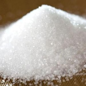 May include: A close-up of a pile of white granulated sugar. The sugar crystals are small and reflect light, creating a sparkling effect. The sugar is on a brown surface.
