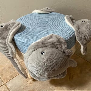May include: A round, light blue ottoman with a woven texture, adorned with plush, gray bunny features. The bunny's head and ears are attached to the ottoman, creating a whimsical and inviting piece of furniture.