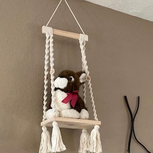 May include: A white macrame hanging shelf with a wooden base and tassels. A brown and white stuffed animal is sitting on the shelf.