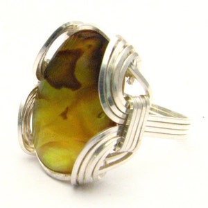 May include: A handcrafted ring featuring a large, oval-shaped, amber-colored stone. The stone is encased in a woven, silver-toned metal setting. The ring's band is a simple, multi-strand design. The stone has brown and yellow hues.