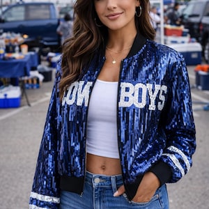 May include: A blue sequined bomber jacket with the text "COWBOYS" in white. The jacket has black trim and white stripes on the sleeves. The person is wearing a white top and denim shorts.