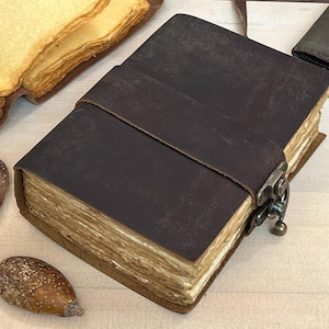 May include: A dark brown leather journal with a metal clasp and strap closure. The journal's pages have a vintage, aged appearance. Decorative keys and other vintage items are arranged around the journal.