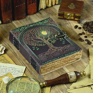 May include: A dark leather-bound journal featuring a tree of life design, with green and gold detailing. The journal is secured with a metal clasp and has aged, textured pages. Other items include a magnifying glass, a wooden box, and coffee beans.