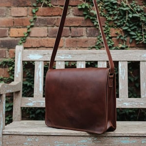 May include: A brown leather messenger bag with a long shoulder strap. The bag is rectangular with a flap closure. The background includes a weathered wooden bench and a brick wall covered in green vines.