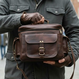 Peut inclure: Un sac messager en cuir marron avec une poignée supérieure, une poche à rabat avant et des poches latérales. Le sac est doté d'une fermeture à boucle et d'une fermeture éclair. Le sac est tenu par une personne portant des gants sans doigts bruns.