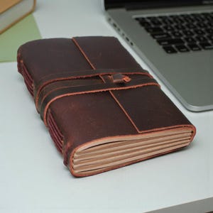May include: A dark brown leather journal with a wrap-around strap closure. The journal's pages are visible, and the cover has a textured appearance. The journal is open, and the pages are a light tan colour. The journal is on a white surface.