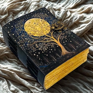 Puede incluir: Un diario decorativo con un diseño celestial. La cubierta es negra con una ilustración dorada de árbol y luna, y páginas doradas. Un cierre de metal asegura el diario, que descansa sobre una tela texturizada.