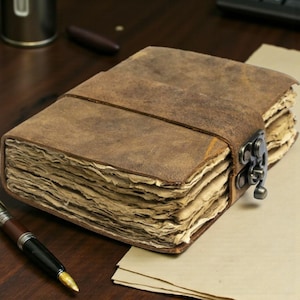 Può includere: Un diario in pelle in stile vintage con chiusura a fermaglio. Il diario ha una copertina in pelle marrone invecchiata e pagine di carta invecchiata. Una penna è appoggiata accanto al diario su una superficie di legno. Il diario è assicurato con un cinturino in pelle.