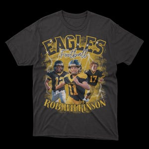May include: A dark grey t-shirt featuring a graphic of three young football players in yellow and black uniforms. The text "Eagles Football" is above the players and "Rob Wilkinson" is below. The background is a stylised lightning bolt design.