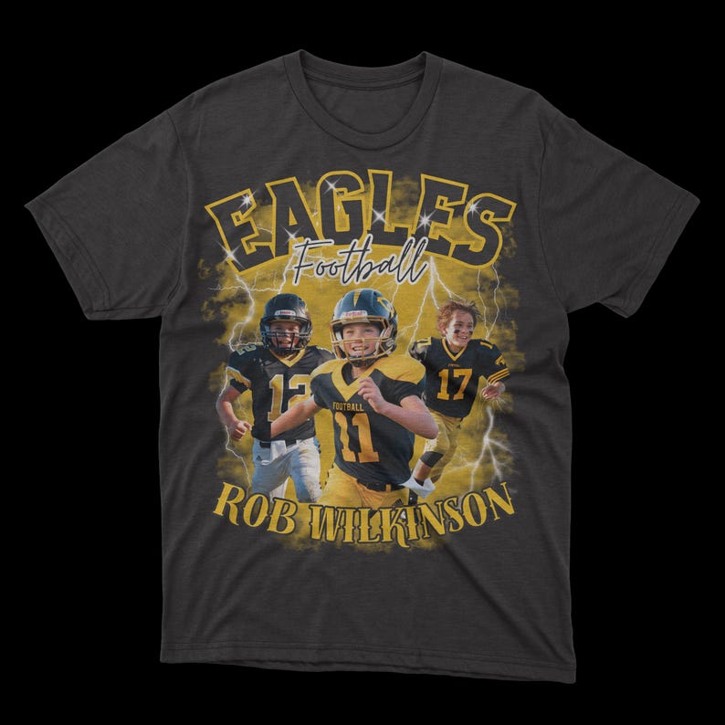 May include: A dark grey t-shirt featuring a graphic of three young football players in yellow and black uniforms. The text "Eagles Football" is above the players and "Rob Wilkinson" is below. The background is a stylised lightning bolt design.