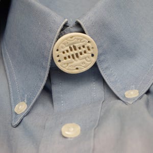 May include: A white, round, decorative button with an intricate carved design. The button is attached to a light blue collared shirt.