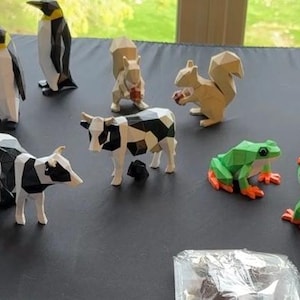 May include: A collection of origami animal figures, including penguins, cows, squirrels, frogs, and elephants. The figures are made of paper and feature geometric designs. The penguins have yellow accents, the cows are black and white, and the frogs are green with orange feet.
