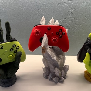 May include: Three unique controller stands. A lime green Xbox controller rests in a black hand-shaped stand. A red controller sits atop a gray crystal-like stand. A black controller is held by a green hand-shaped stand.