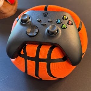 May include: A black wireless video game controller resting on top of a textured orange and black basketball. The controller features a directional pad, two analog sticks, and four primary action buttons labeled with X, Y, A, and B symbols.