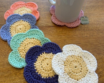 May include: Six crocheted flower-shaped coasters in various colors, including pink, blue, green, purple, and white. The coasters are arranged in a row on a wooden surface. A white mug with a tea bag is sitting on a pink coaster.