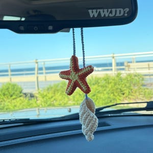 May include: A hanging car ornament featuring a crocheted orange and yellow starfish paired with a cream-colored crocheted seashell, suspended by a light gray cord from a rearview mirror that displays the white text WWJD? on its lower edge.