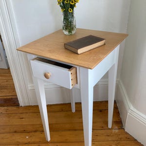 May include: White nightstand with a single drawer and a wooden top. The drawer has a wooden knob and is partially open. A book is on top of the nightstand.