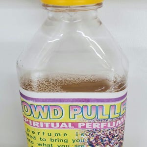 May include: A clear plastic bottle with a yellow cap filled with a light brown liquid. The bottle has a label that reads "Crowd Puller Spiritual Perfume" and includes a picture of a crowd of people. The label also includes the text "Perfume is created to bring you into what you are. It is good for Ifa and everybody. Direction: Sprinkle this perfume 3 times in your place of work early in the morning. Spiritual Products."