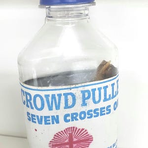 May include: A clear plastic bottle with a blue cap filled with a blue liquid. The label reads "Crowd Puller Seven Crosses Oil" with a red cross inside a circle and the numbers "121, 23, 41". The label also reads "Alfa Ele Eli OSD Pro".