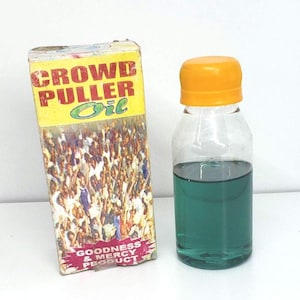 May include: A clear glass bottle with a yellow cap, filled with teal liquid, next to a yellow box. The box reads "CROWD PULLER Oil" and "GOODNESS & MERCY PRODUCT". The box has an image of a crowd.