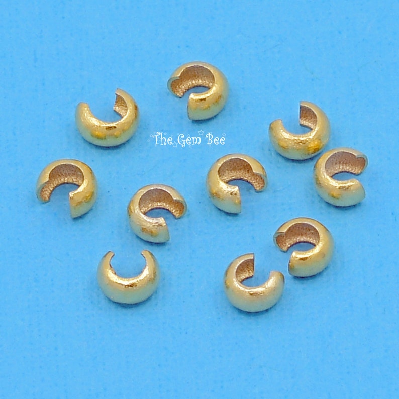 May include: Ten gold-coloured crimp beads, used for jewellery making, are scattered on a blue background. The crimp beads are small, round, and have a hole in the centre.