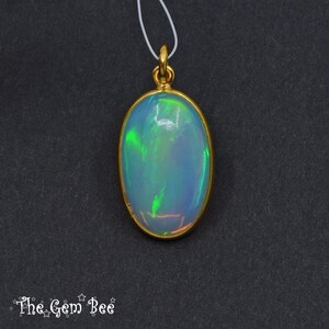 May include: Oval opal pendant with a gold-coloured bezel and bail. The opal displays iridescent colours of green, blue, and orange. The pendant hangs from a thin white cord against a dark background. The Gem Bee logo is in the bottom left corner.