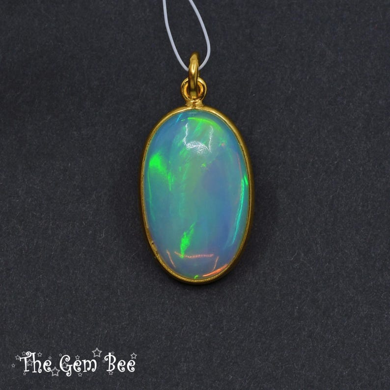 May include: Oval opal pendant with a gold-coloured bezel and bail. The opal displays iridescent colours of green, blue, and orange. The pendant hangs from a thin white cord against a dark background. The Gem Bee logo is in the bottom left corner.