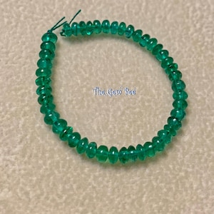 Peut inclure: Un fil de perles de pierres précieuses vert vif, probablement des émeraudes. Les perles sont petites, rondes et polies, reflétant la lumière. Le texte "The Gem Bee" est visible sur la surface. Les perles sont enfilées sur un fil vert, formant un bracelet ou un collier.