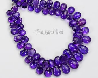 Puede incluir: Un hilo de piedras preciosas de amatista de color púrpura intenso en forma de pera, dispuestas en forma de U con una borla de color púrpura oscuro en la parte superior. El texto The Gem Bee está impreso en el centro. Cada piedra es lisa y translúcida.