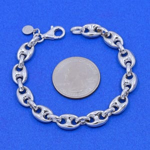 May include: A silver-colored chain bracelet with a lobster clasp, featuring a series of oval links. A U.S. quarter coin is placed next to the bracelet for size comparison. The bracelet is displayed on a blue background.