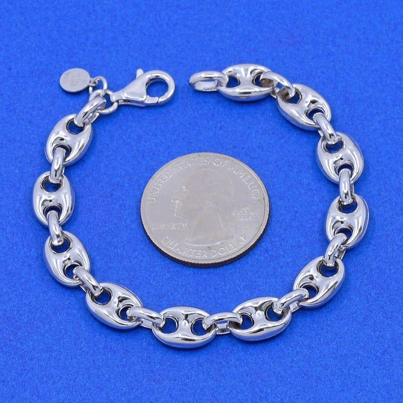 May include: A silver-colored chain bracelet with a lobster clasp, featuring a series of oval links. A U.S. quarter coin is placed next to the bracelet for size comparison. The bracelet is displayed on a blue background.