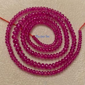 Puede incluir: Un largo hilo de pequeñas cuentas redondas y translúcidas de color rosa fucsia dispuestas en espiral. Las cuentas están ensartadas en un hilo rojo fino. El texto The Gem Bee está impreso en azul en el centro de la espiral de cuentas.