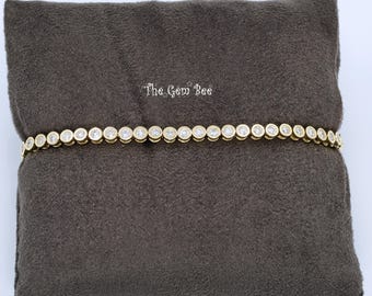 Puede incluir: Una pulsera de tenis en tono dorado con una fila continua de piedras redondas transparentes engastadas en bisel. La pulsera se muestra horizontalmente en el centro. El texto "The Gem Bee" está impreso en blanco sobre el centro de la pulsera.