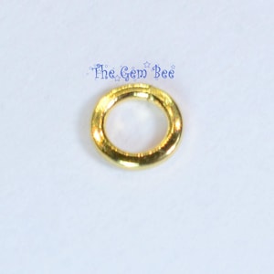 May include: A small, circular gold-toned jump ring with a smooth, polished finish. The metal ring features a single visible seam where the ends meet. Above the ring, the text The Gem Bee is printed in blue with small star accents.