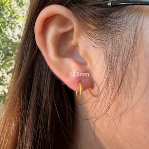 Puede incluir: Un peque&ntilde;o pendiente de aro dorado con textura trenzada, que presenta un di&aacute;metro de 12,5 mm. El pendiente se lleva en el l&oacute;bulo de la oreja y tiene un acabado met&aacute;lico pulido que refleja la luz, mostrando un dise&ntilde;o circular sencillo.