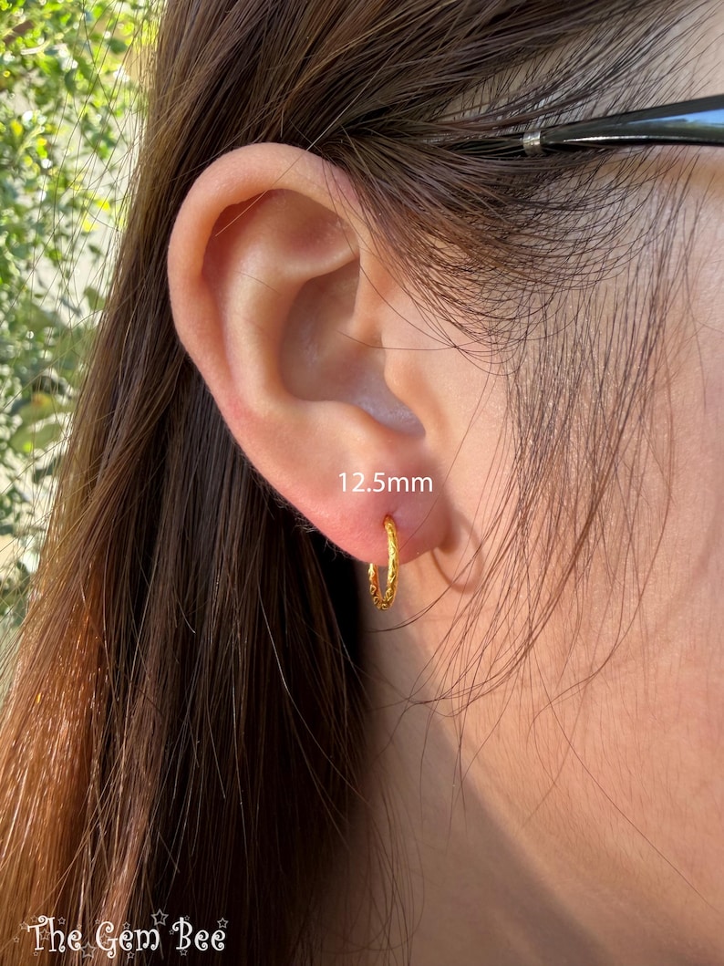 Puede incluir: Un peque&ntilde;o pendiente de aro dorado con textura trenzada, que presenta un di&aacute;metro de 12,5 mm. El pendiente se lleva en el l&oacute;bulo de la oreja y tiene un acabado met&aacute;lico pulido que refleja la luz, mostrando un dise&ntilde;o circular sencillo.