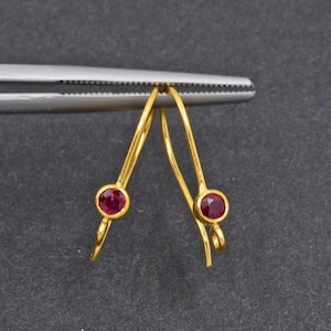 Op de afbeelding: Een paar goudkleurige oorbellen met een kleine, ronde, rode edelsteen aan de basis van elke oorbel. De oorbellen worden vastgehouden door een zilveren gereedschap tegen een donkergrijze achtergrond.