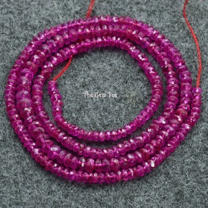 May include: A strand of pink ruby beads, approximately 3mm in size, with a faceted, slightly elongated shape. The beads are strung on a red thread and are arranged in a circular pattern. The text "The Gem Bee" is visible in the image.