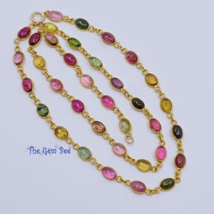 Op de afbeelding: Een gouden ketting met ovale edelstenen in verschillende kleuren, waaronder roze, groen, geel en bruin. De edelstenen zijn verbonden door een fijne gouden ketting. Het logo The Gem Bee is zichtbaar.