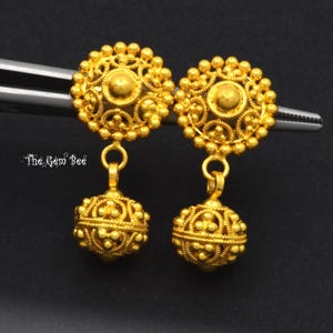May include: A pair of gold-toned drop earrings featuring circular studs with intricate beaded filigree patterns and matching spherical dangles. The earrings are held by silver-toned tweezers. The text The Gem Bee is visible on the left side.
