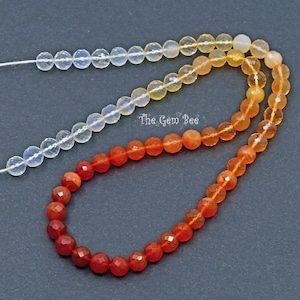May include: A strand of faceted gemstone beads in a gradient of colors, from clear to orange to red. The beads are strung on a white thread. The text "The Gem Bee" is visible in the image.