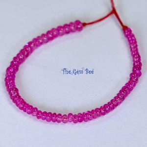 Op de afbeelding: Een streng van kleine, ronde, felroze kralen geregen aan een dun rood koord. De kralen zijn uniform van grootte en vorm en vormen een doorlopende cirkelvormige lus. De tekst The Gem Bee is in het blauw in het midden afgedrukt.