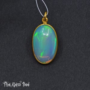 May include: Oval opal pendant with a gold-coloured bezel and bail. The opal displays a play of colour with hues of blue, green, and orange. The pendant hangs from a thin white cord against a dark background. The Gem Bee logo is in the bottom left corner.