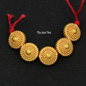 May include: Five gold-colored round beads with a textured surface, strung on a red cord. The beads have a raised center and a circular pattern of small dots around the edge. The text "The Gem Bee" is visible in the background.