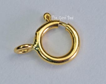May include: A gold-toned circular spring ring clasp featuring a small lever mechanism and an attached jump ring. The metal surface is smooth and reflective. The text The Gem Bee is visible in the upper right area of the jewelry component.