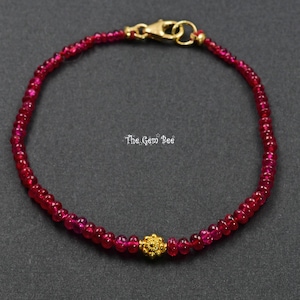 May include: A beaded bracelet featuring small, faceted, deep red gemstone beads arranged in a circular pattern. A single, intricate, gold-toned floral-shaped bead is centered at the bottom. The bracelet includes a gold-toned lobster clasp closure.