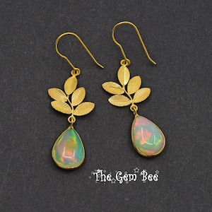 Può includere: Un paio di orecchini color oro con un design a foglie e pietre preziose di opale a forma di goccia. Gli opali mostrano un mix di colori, tra cui verde, rosa e giallo. Gli orecchini sono su uno sfondo scuro.