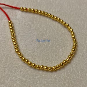 May include: A strand of small, polished gold-colored spherical beads arranged in a curved line. The beads are strung on a thin red cord, with the text The Gem Bee visible in blue lettering near the center of the curved bead arrangement.